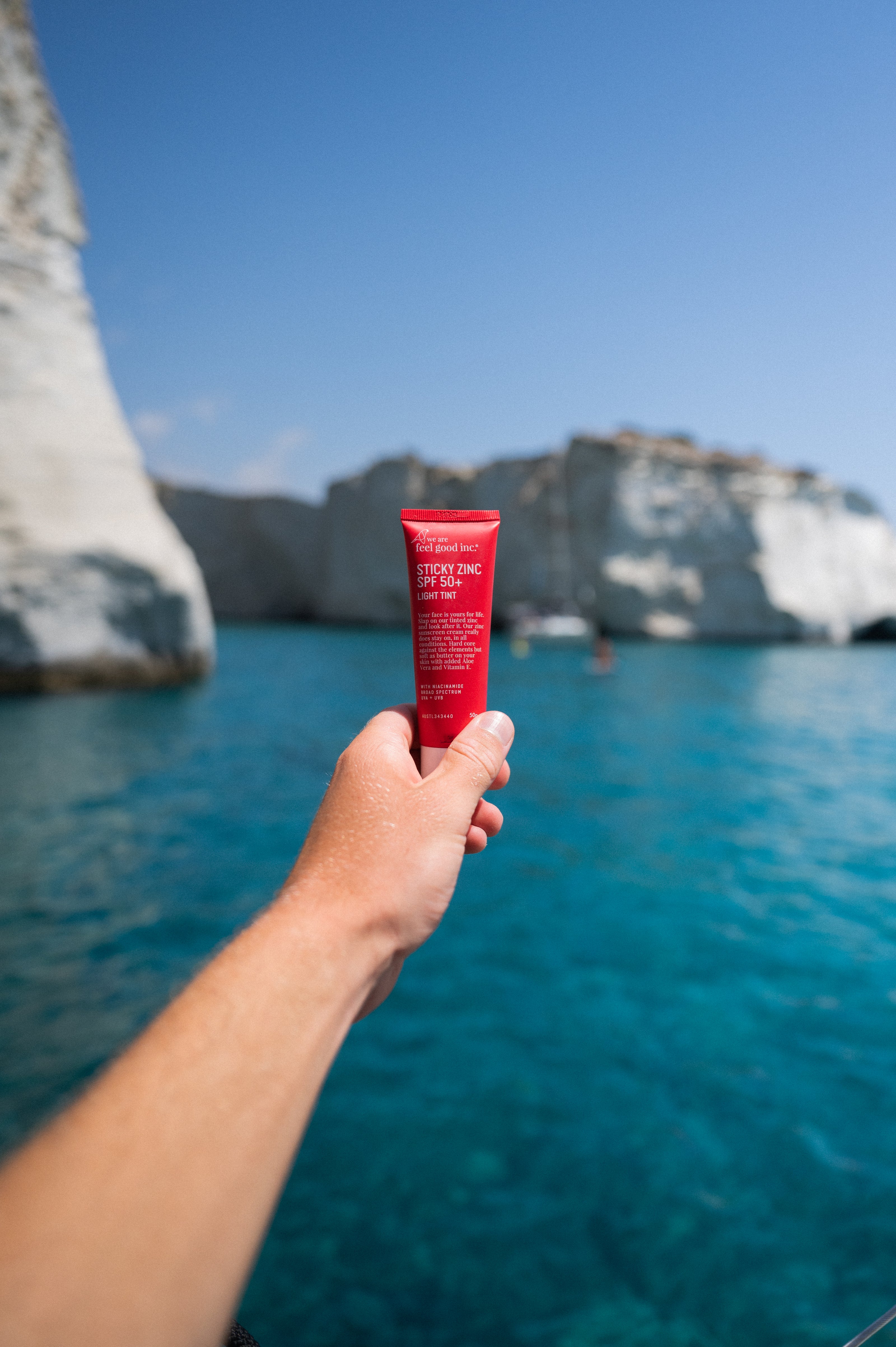 A hand holding a plastic tube of We Are Feel Good Inc. Sticky Zinc SPF50+ sunscreen in front of clear blue waters with white limestone cliffs in the distance.