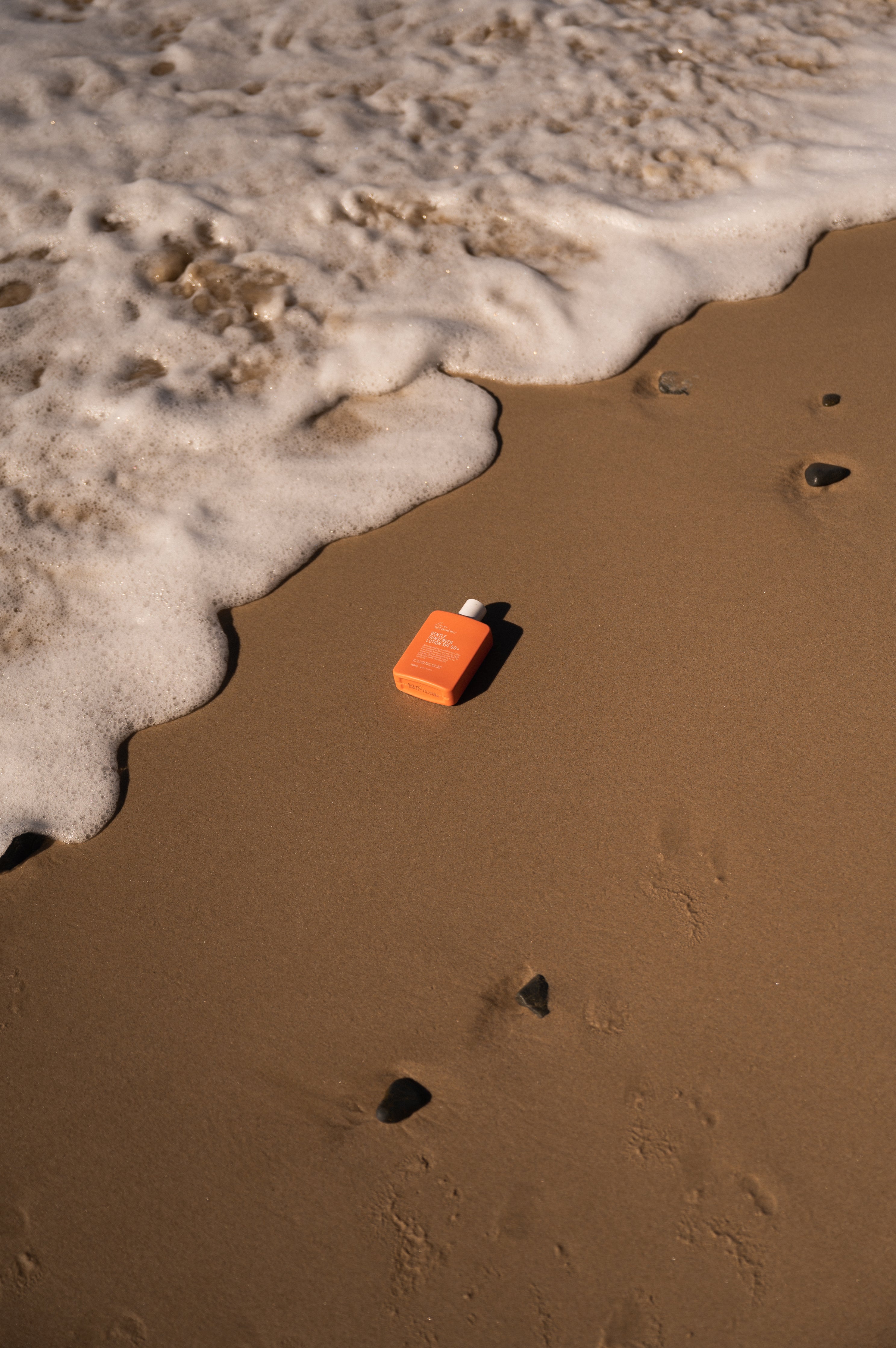 We Are Feel Good Inc. Sensitive Sunscreen is placed on wet sand, with the foamy edge of a wave almost touching it, under warm sunlight. Footprints and small stones mark the sand around it.